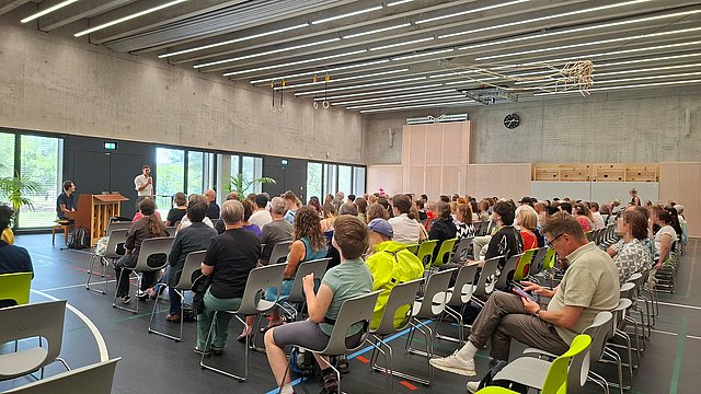 Viele Menschen sitzen in Stuhlreihen hintereinander in der Sporthalle. Vor ihnen sitzt ein Mann an einem Klavier. Daneben spricht der Schulleiter in ein Mikrofon